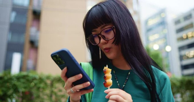 Japanese woman, snack and typing with phone for social media, mobile notification and dango in city park. Commuter, smartphone and search online for travel break, location or urban street of Tokyo