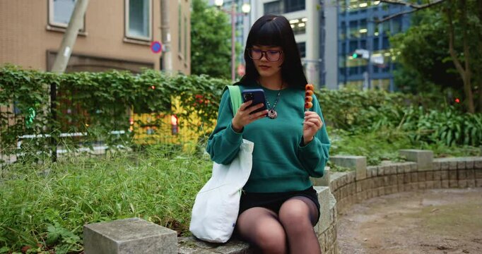 Japanese woman, park and typing with smartphone for social media, mobile notification and dango snack in city. Commuter, phone and search online for travel break, location or urban street of Tokyo