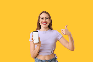 Young woman with mobile phone showing thumb-up gesture on yellow background