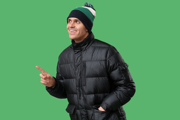 Young man in stylish puffer jacket and hat pointing at something on green background