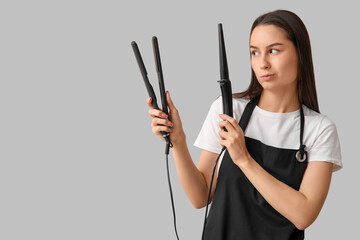 Female hairdresser with curling iron and flattening iron on grey background