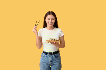 Beautiful young Asian woman holding tasty sushi rolls on wooden tray and chopsticks on yellow...