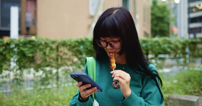 Japanese woman, snack and typing with smartphone for social media, mobile notification and dango in city park. Commuter, phone and search online for travel break, treat or urban street of Tokyo