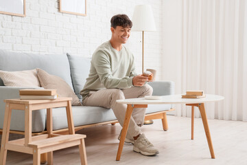 Young man with cup of tea taking tasty muffin from table at home