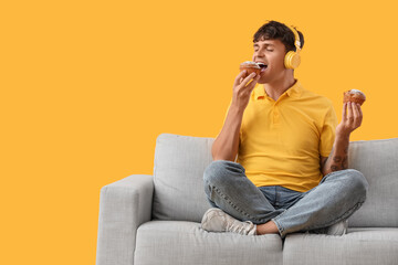 Young man in headphones eating tasty muffin on sofa against yellow background