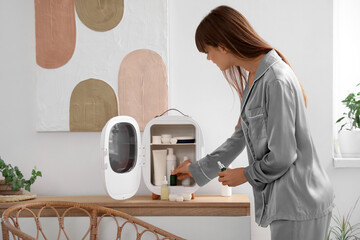 Beautiful young woman taking cosmetics from refrigerator at home