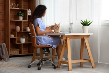 Beautiful young happy African-American woman working with laptop, cup of coffee and her cute Maine Coon cat at home