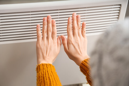 Young woman warming hands near electric convector heater at home, closeup