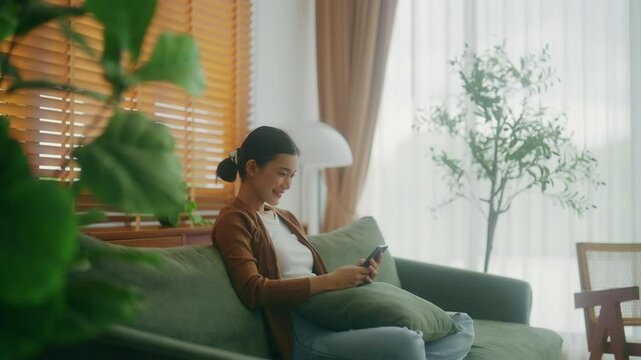 Smiling pretty young Asian woman sitting on cozy couch, using funny mobile apps in living room. Woman at home, Doing online shopping, Messaging friends, Posting on social Media