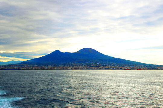Crisp view from Naples at the staggering Mount Vesuvius soaring in the dramatic-looking sky full of clouds pierced by diffuse sunlight, swathes of buildings under it, and the Tyrrhenian Sea