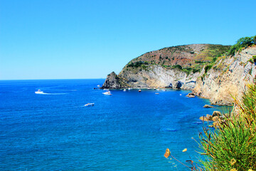 Pristine scenery from Ischia with the rocky formations at Punta Chiarito, beautiful deep blue Tyrrhenian Sea, white yachts, boats, wakes, emerald shades and greenery under a gradient azure sky