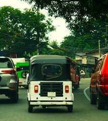 Tuk Tuk jedzie ruchliwą drogą. Ruch drogowy, liczne samochody poruszają się po drodze. Mały Tuk Tuk widoczny z tyłu. Typowy pojazd używany na Filipinach.