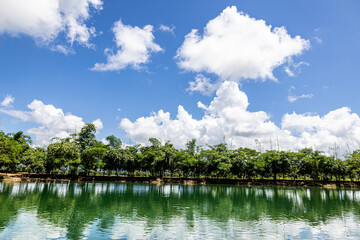 White clouds float in the sky, reflecting beautifully on the water's surface. Trees line the shore, creating a peaceful atmosphere. This photo was taken in Karen State, Myanmar in August.