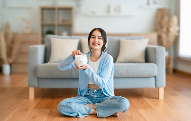 Young Asian woman holding piggy bank and sitting on the floor at home. Save money and financial...
