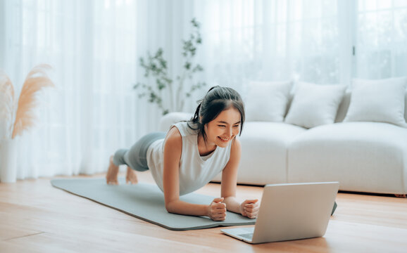 Young Asian woman in sportswear doing plank on training mat while watching online workout tutorials on laptop in the living room. Sport and recreation concept