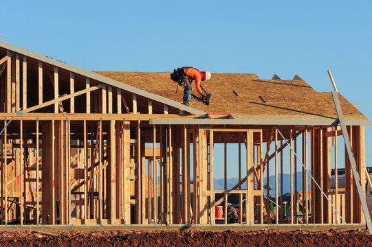 A construction worker atop a tract home under construction in a new subdivision, wearing a tool belt, a hard hat, and fall protection equipment
