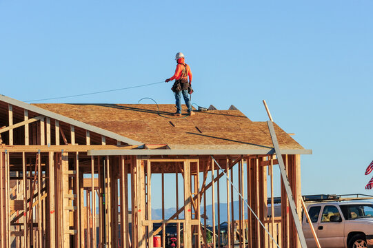 A construction worker atop the structure of a home under construction in a new subdivision, wearing a tool belt, a hard hat, and fall protection equipment