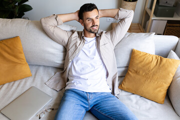 Man relaxing on the sofa with hands behind the head looking away after working with loptop. Feeling satisfaction