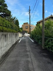 narrow street in a suburban Japanese neighborhood - one-way road between a concrete wall and a fence, with telephone poles and wires, houses, bushes and trees and greenery, beneath a blue summer sky