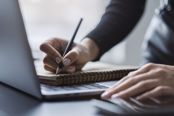 Close perspective of a woman's hand scribbling in a notepad on a stylish laptop, with a soft blur...