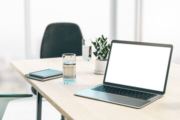 Perspective view on blank white laptop screen on light wooden table in sunny office with glass of water, notebooks and flowerpot on black chair and big blurred window background. 3D rendering, mock up
