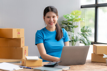 Smiling Business Owner:  A young woman smiles confidently at the camera while working on her laptop at her home office, surrounded by parcels ready for shipping. This image evokes a sense of success.