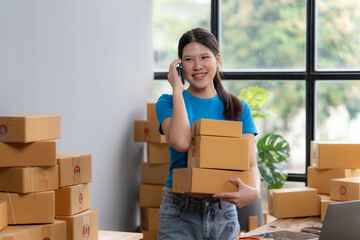 Online Business Success: A young, smiling Asian woman entrepreneur confidently handles a stack of shipping boxes while talking on her phone, surrounded by inventory in a home office setting.  