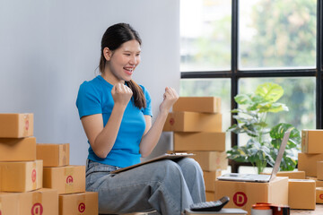 Online Business Success: Happy young Asian woman celebrates her e-commerce success surrounded by boxes, laptop, and clipboard.  