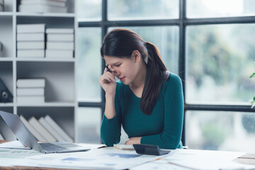 Overwhelmed Businesswoman: A young Asian businesswoman sits at her desk, her hand to her forehead, clearly stressed and overwhelmed by paperwork and deadlines.