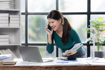 Obraz premium Confident and Connected: A woman in a green top exudes professionalism and positivity as she takes a call while multitasking at her office desk, showcasing a modern, organized workspace.