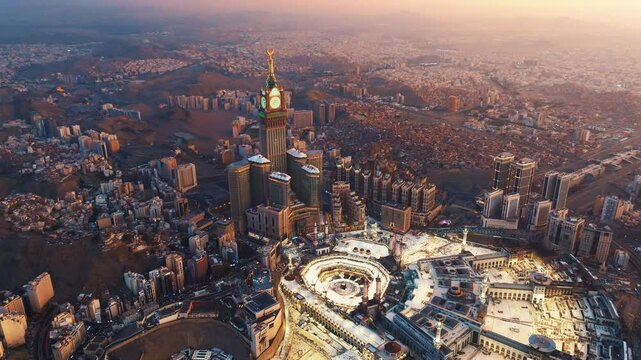 Aerial view of the illuminated Abraj Al-Bait Clock Tower and Grand Mosque in Mecca at dawn, surrounded by the sprawling city and mountains bathed in soft morning light