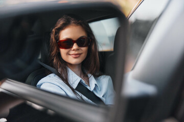 Woman, sunglasses, car a woman wearing sunglasses smiles while sitting in the rear view mirror of a car, her hand gently resting on the side of her face, conveying a relaxed and confident mood