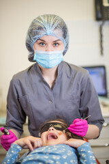 In a dental clinic, a young girl wears protective sunglasses as she sits in the dentist's chair, while a smiling dental assistant prepares for her treatment with care and focus.