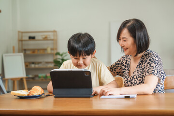 A mother teaches her son to study online at home, using a digital tablet for learning. They sit together at a table with snacks, promoting homeschooling and family bonding.