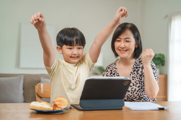 A mother teaches her son to study online at home, using a digital tablet for learning. They sit together at a table with snacks, promoting homeschooling and family bonding.