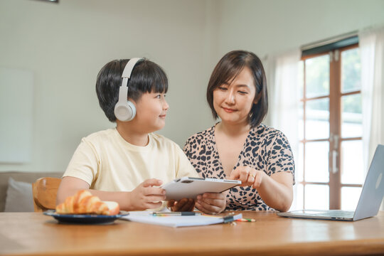 A mother teaches her son to study online at home, using a digital tablet for learning. They sit together at a table with snacks, promoting homeschooling and family bonding.
