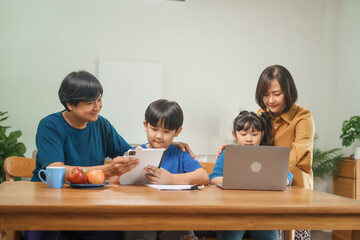 Asian parents and their children sit together at the table in the morning, studying online using tablets and computers, fostering a productive and focused family learning environment
