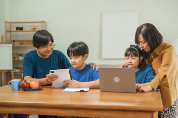 Asian parents and their children sit together at the table in the morning, studying online using tablets and computers, fostering a productive and focused family learning environment