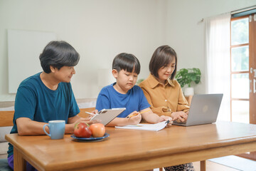 Fototapeta premium Asian parents and their children sit together at the table in the morning, studying online using tablets and computers, fostering a productive and focused family learning environment