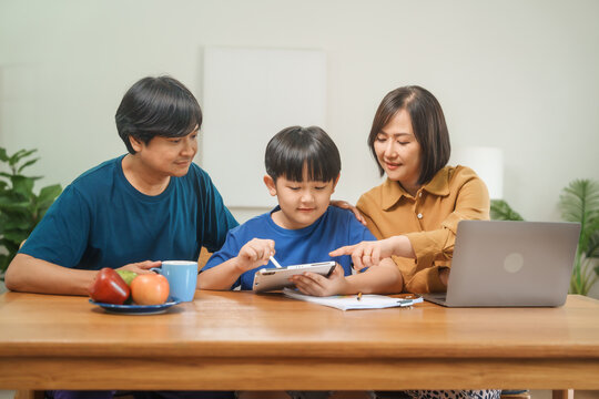 Asian parents and their children sit together at the table in the morning, studying online using tablets and computers, fostering a productive and focused family learning environment