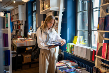 Interested focused middle aged woman spending free leisure time choosing book shopping in bookstore. Thoughtful mature female with book in hands standing near bookcase in library, enjoying reading.