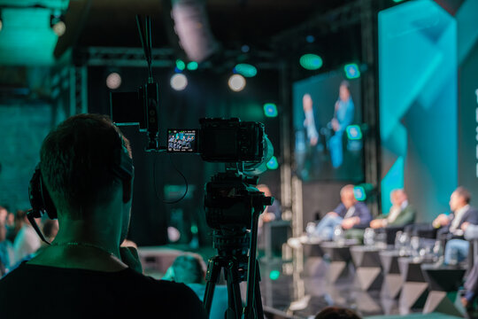 Close-up of professional video camera capturing speakers on stage during business conference. Focus on technology and communication in live event filming.
