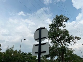 A blank road sign stands against a cloudy sky, surrounded by trees and streetlights.