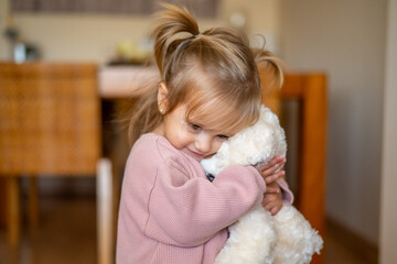 Happy Child Holding A Teddy Bear In A Warm Living Room, Radiating Love And Security.Loving Baby Girl Hugging A Plush Bear In A Family Setting, Symbolizing Care And Childhood Comfort  