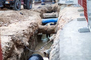 A construction site showing a trench with a blue pipe being installed and a worker performing...