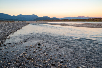 The Tagliamento river riverbed at dusk with, rocks and pebbles and sun rays reflections on the waters with the carnic prealps in the background