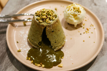close up of dessert pistachio lava cake with syrup lava chocolate cream coming out from the cake with vanilla ice cream on the side and nuts as toppings with two spoons on the plate