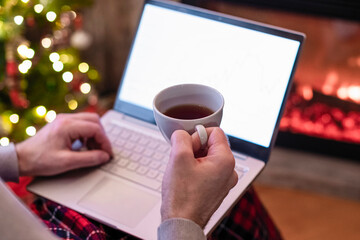 Christmas. Man with laptop sitting near christmas tree and fireplace with cup of tea. Concept of online service in holidays. Back view.