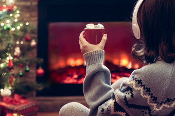 Woman in headphones with cup of hot cocoa and marshmallow sitting and warming at winter evening near fireplace flame and  christmas tree.