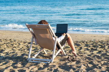 Back view of alone young brunette woman freelancer in office clothes with a laptop looking on the sea sitting on the deck chair on beach. Summer concept